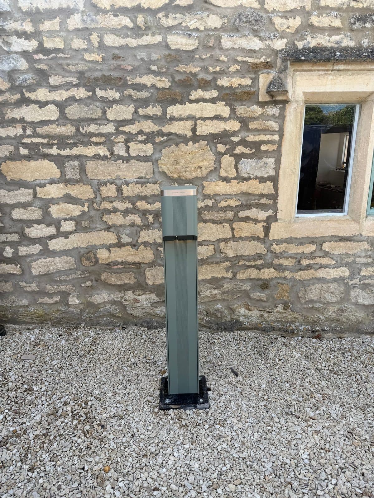 Sage-green EV charging pedestal bolted to a gravel driveway beside a stone cottage