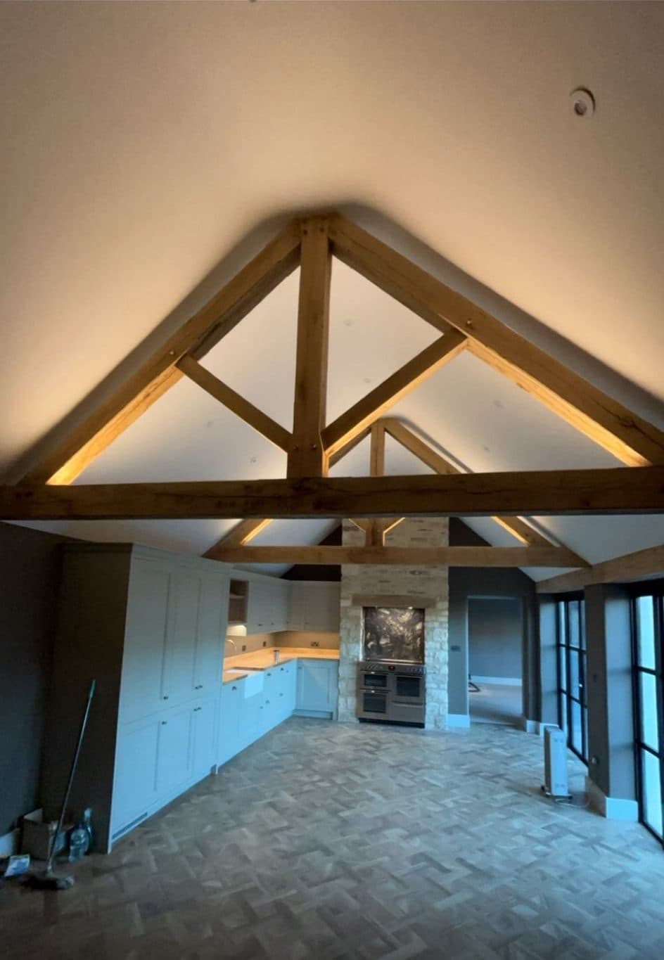 Open-plan barn kitchen and living area with uplit oak king-post trusses and a stone chimney breast